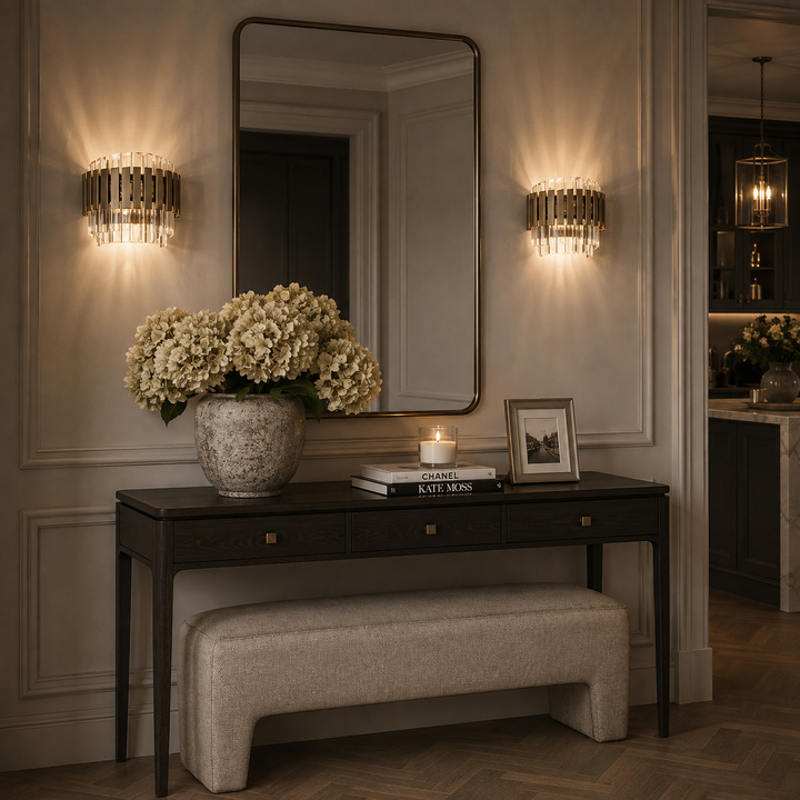 A styled interior scene with a dark wood console table, upholstered bench, large wall mirror, and two illuminated cylindrical wall sconces. A vase of cream flowers, books, and framed art sit on the table against a paneled wall.