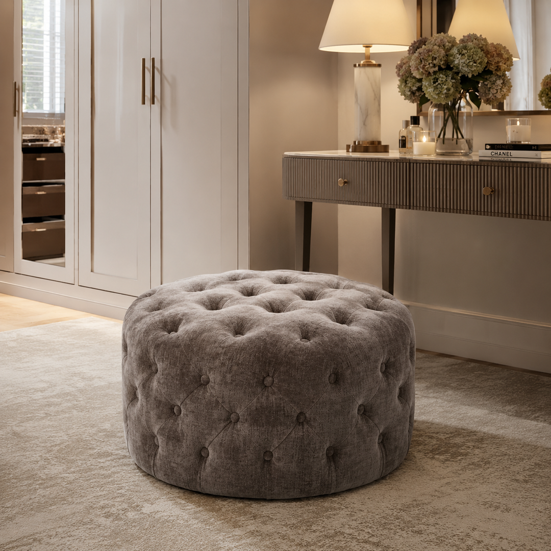A round tufted ottoman in textured gray fabric, positioned on a light beige rug in a styled home office or dressing room setting with a console table and lamp in the background.