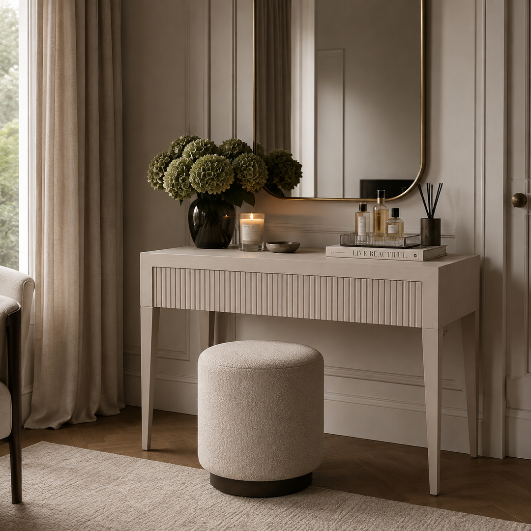 A cream-colored vanity table with a fluted front and tapered legs, styled in a warm, neutral-toned room with a matching round ottoman, a large mirror, and decorative items including a vase of green hydrangeas and candles.