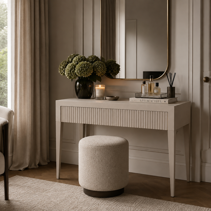 A cream-colored vanity table with a fluted front and tapered legs, styled in a warm, neutral-toned room with a matching round ottoman, a large mirror, and decorative items including a vase of green hydrangeas and candles.