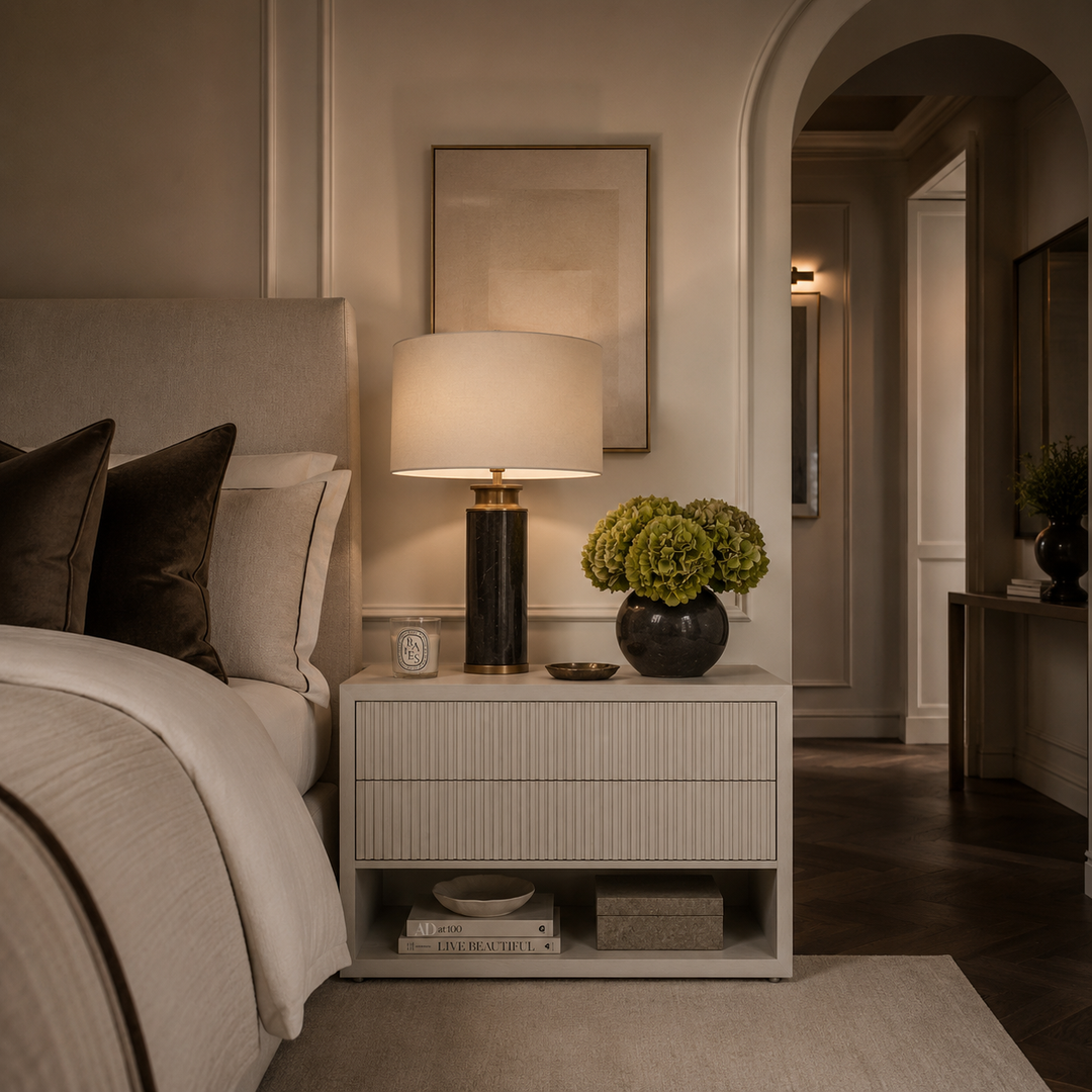 A modern bedroom scene featuring a white ribbed nightstand with a lit table lamp, a vase of green hydrangeas, and a small clock, set next to a bed with neutral-toned bedding.