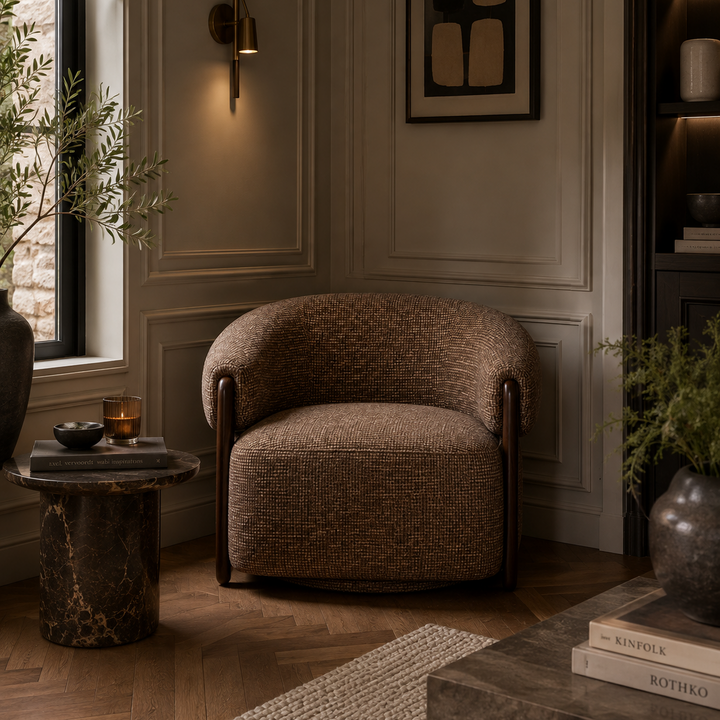 A cozy upholstered armchair in a textured brown fabric sits in a styled interior room with wood paneling, a wall sconce, framed abstract art, a side table, and plants.