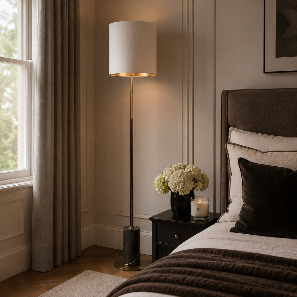A modern floor lamp with a white drum shade and a slim gold-finished metal stand, styled in a warm, elegant bedroom next to a bed with white and dark brown bedding.