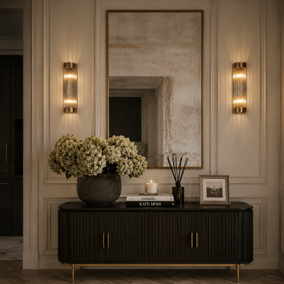A styled interior entryway with a dark wood console cabinet, a large floral arrangement, a mirror, and two matching wall sconces mounted on a paneled beige wall.