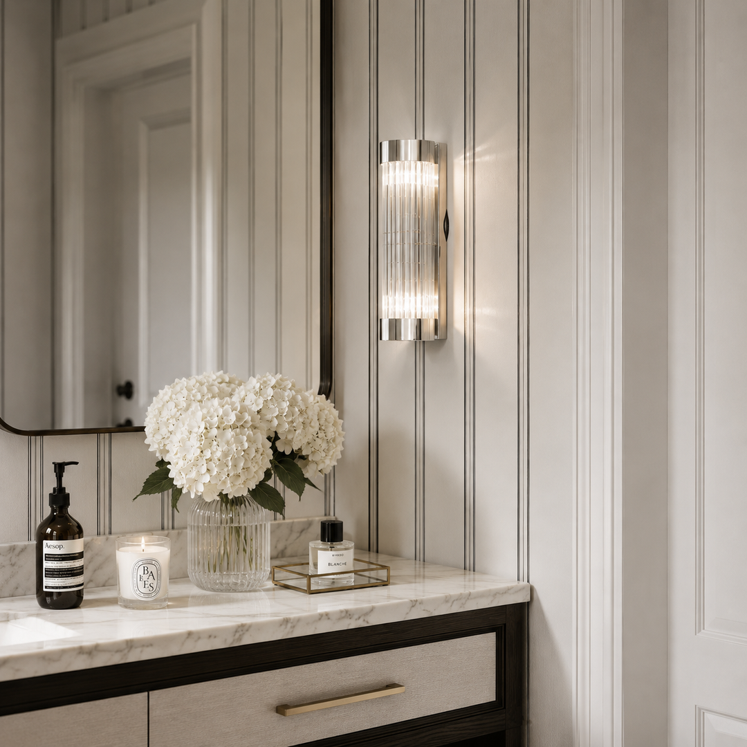 A modern wall-mounted sconce with a vertical ribbed glass shade and brushed metal frame, glowing with warm light on a paneled wall above a bathroom vanity.