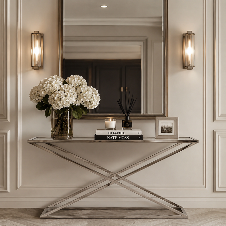 A modern console table with a mirrored glass top and an X-shaped brushed metal base, styled in a classic entryway. The table is decorated with a large vase of white hydrangeas, a candle, a reed diffuser, and a small framed print.
