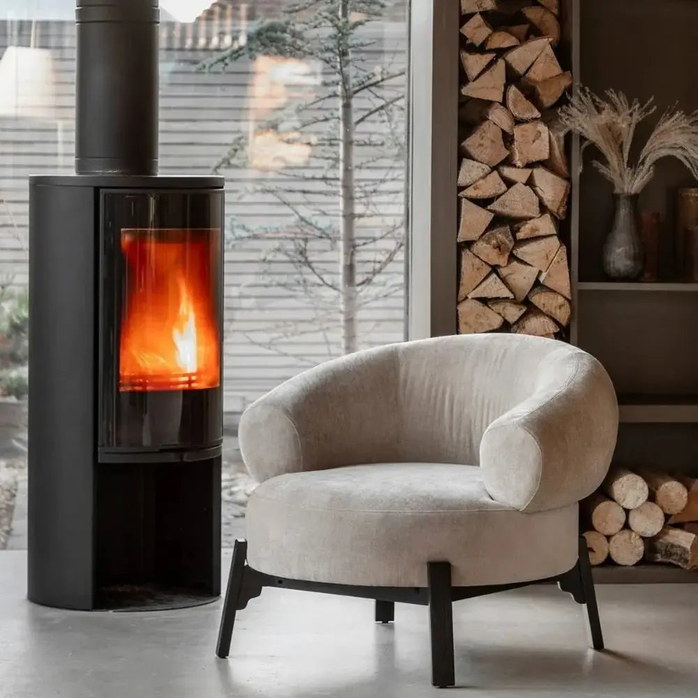 Modern armchair in a room with a lit stove and stacked firewood.