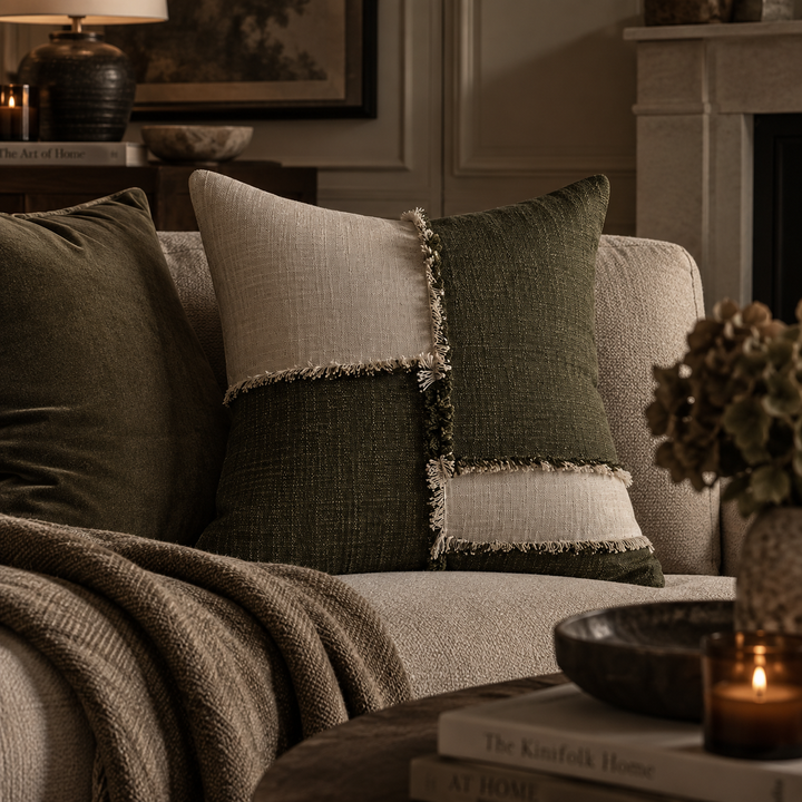 A styled home interior scene showing a patchwork throw pillow on a beige sofa, with matching olive green pillows and a knit throw blanket in a cozy room setting.
