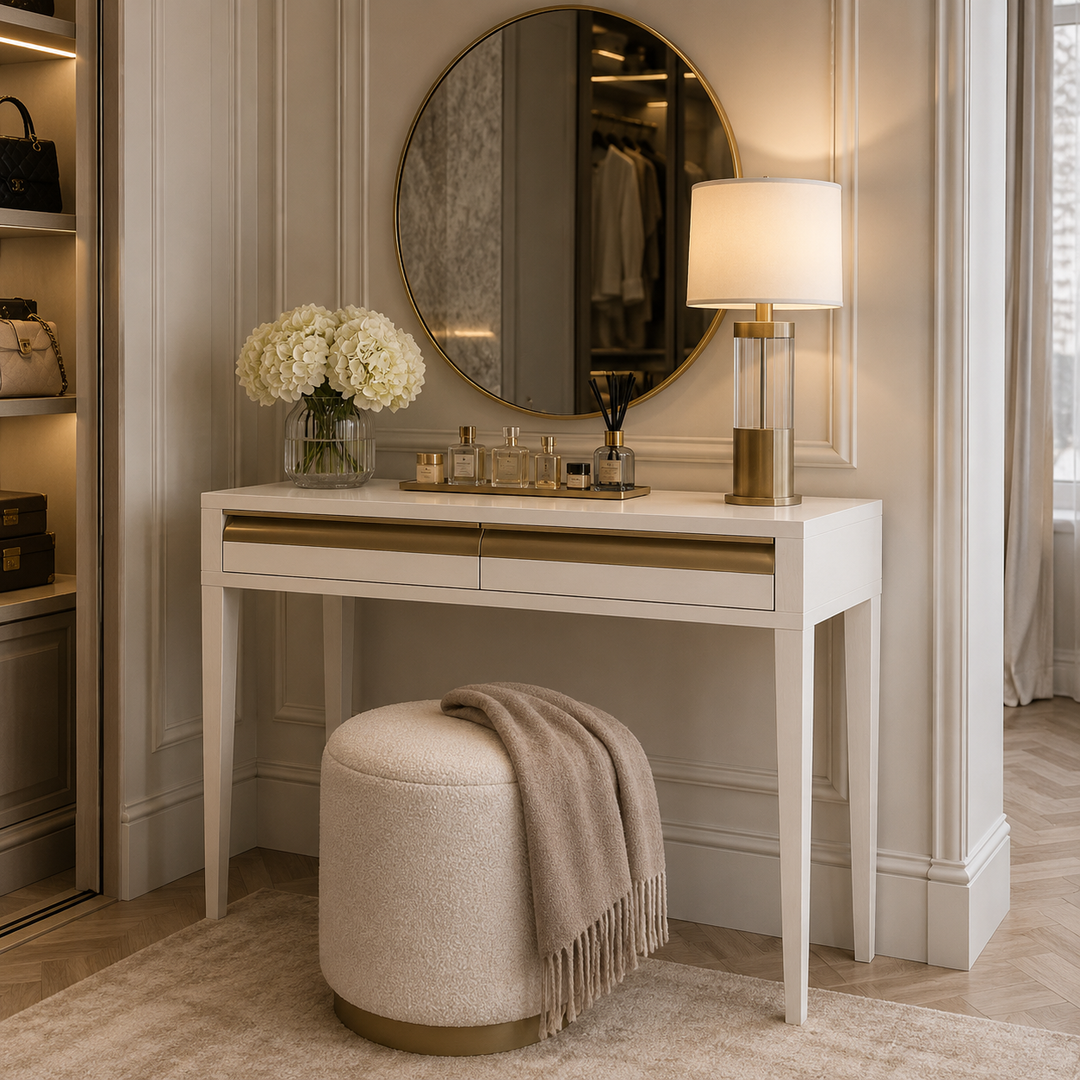 A white vanity desk with gold trim and drawers, styled in a luxurious room with a round mirror, a lit table lamp, a beige ottoman with a fringed throw blanket, and a vase of white flowers.