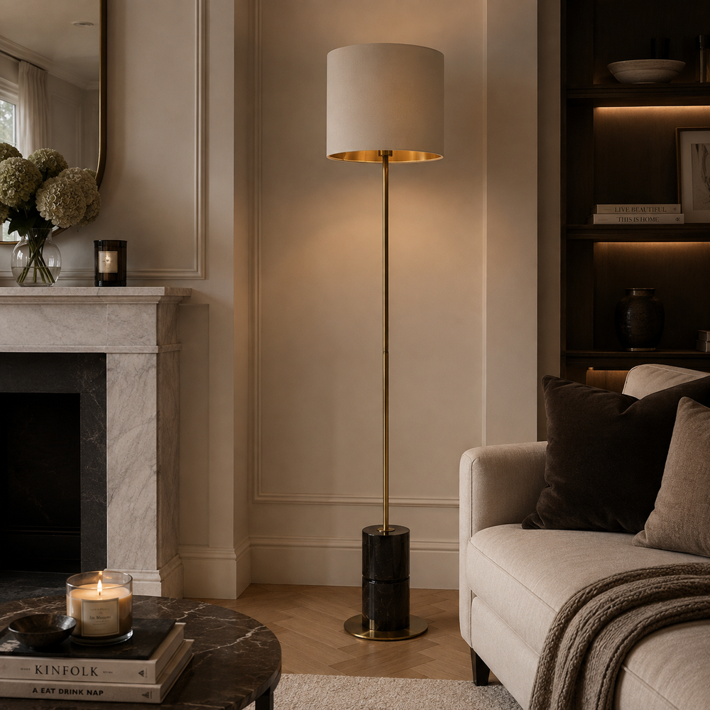 A modern floor lamp with a cylindrical white drum shade, a brushed gold metal top rim, and a slim gold stand with a black cylindrical base, styled in a warm, elegant living room next to a marble fireplace and a beige sofa.