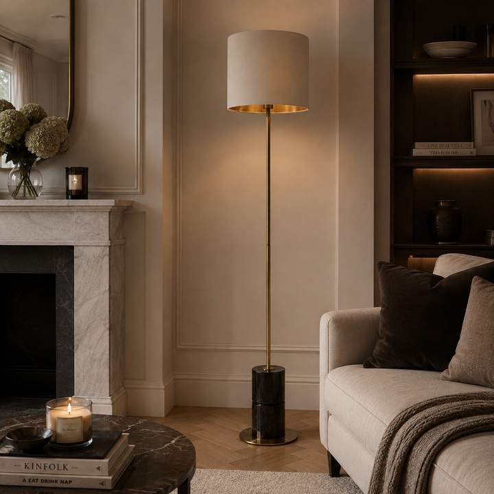 A modern floor lamp with a cylindrical white drum shade, a brushed gold metal top rim, and a slim gold stand with a black cylindrical base, styled in a warm, elegant living room next to a marble fireplace and a beige sofa.