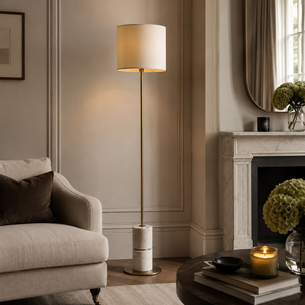 A tall floor lamp with a warm white drum shade and a slim brass-colored pole stands in a styled living room next to a beige armchair and in front of a fireplace.