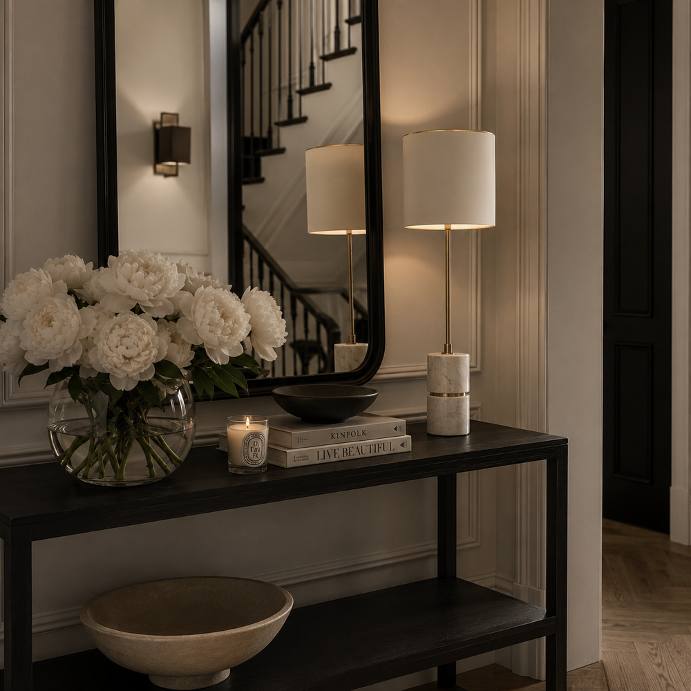 A modern table lamp with a white cylindrical base and a gold-toned stem stands on a dark console table in a styled entryway. A large mirror behind it reflects the lamp, and the scene includes a vase of white flowers, books, a candle, and bowls.