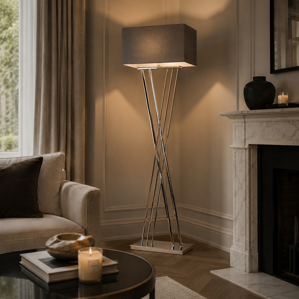 A modern floor lamp with a dark gray fabric drum shade and three intersecting chrome metal legs, styled in a warm, elegant living room next to a fireplace and sofa.