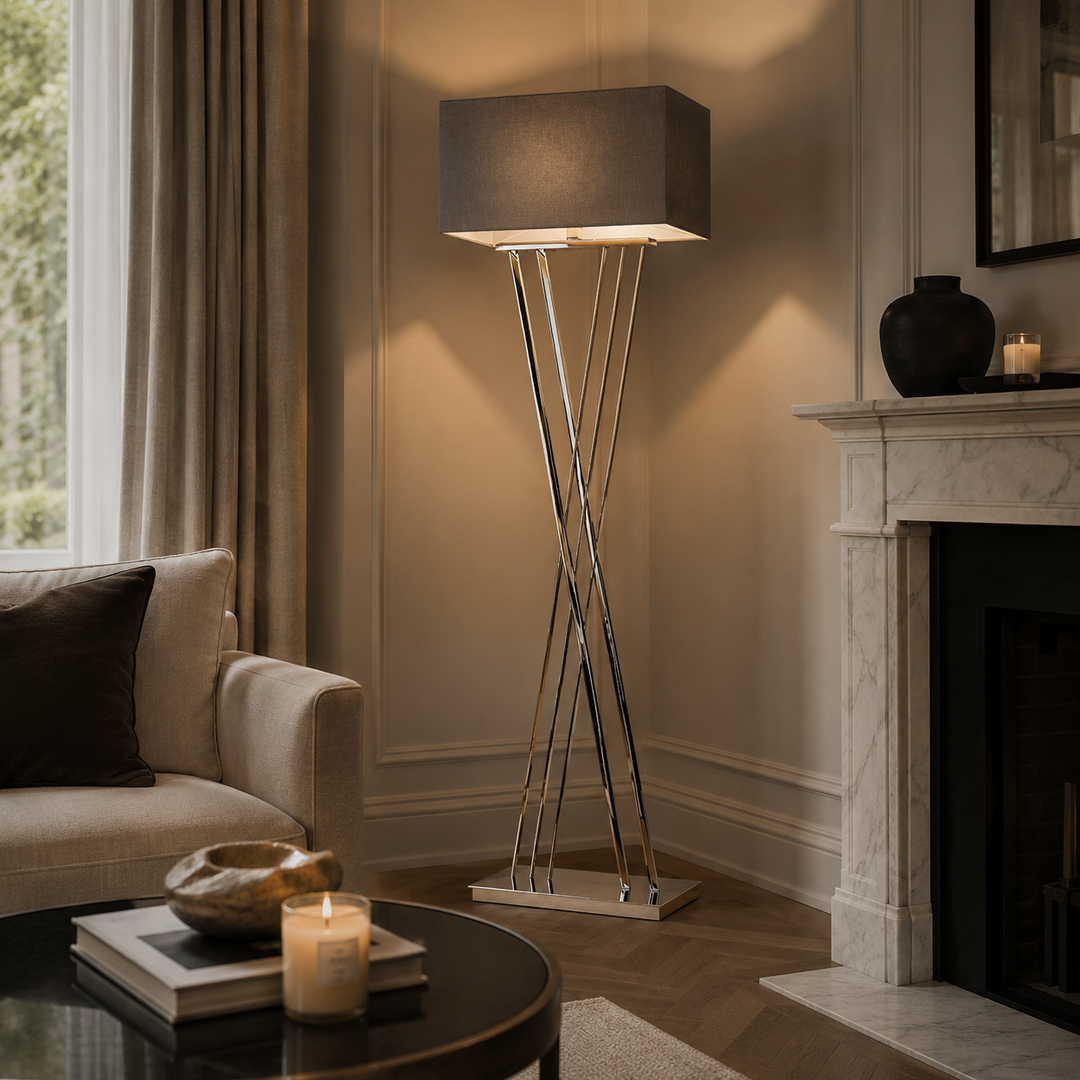 A modern floor lamp with a dark gray fabric drum shade and three intersecting chrome metal legs, styled in a warm, elegant living room next to a fireplace and sofa.