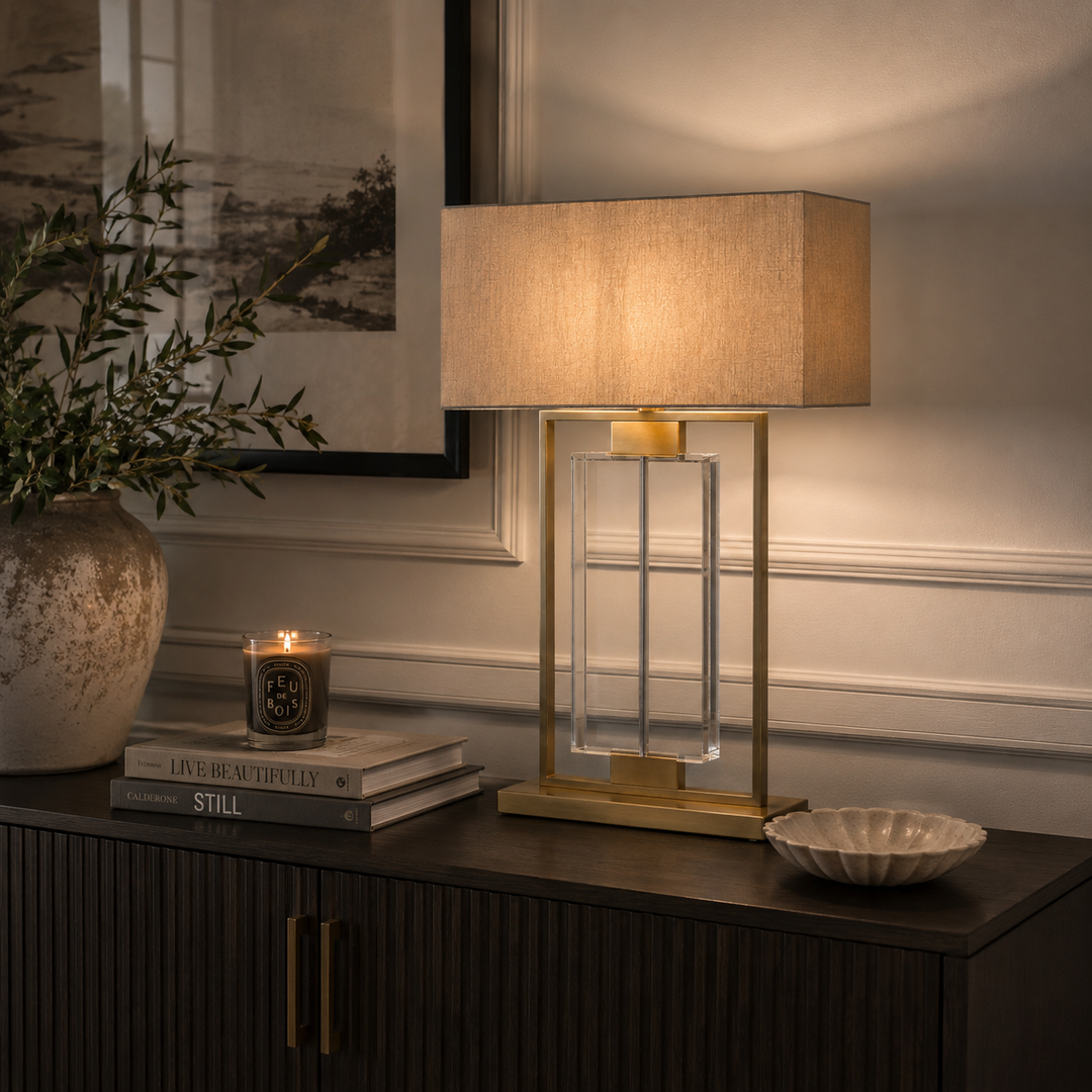 A modern table lamp with a warm beige fabric drum shade, a clear glass rectangular column, and a gold metal frame, shown lit on a dark wood console table in a styled interior setting.