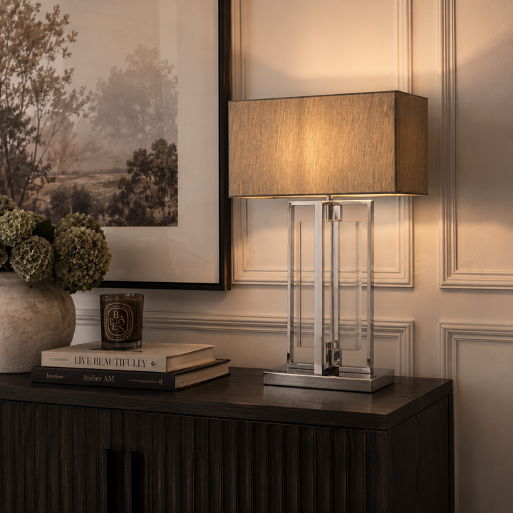 A table lamp with a warm beige fabric drum shade and a clear glass column on a square silver base, styled on a dark wood sideboard in a decorated interior setting.