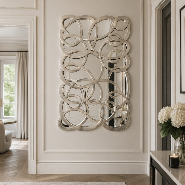An ornate rectangular wall mirror with an intricate, interlocking silver loop pattern, mounted on a cream-colored paneled wall in a beautifully styled interior hallway.