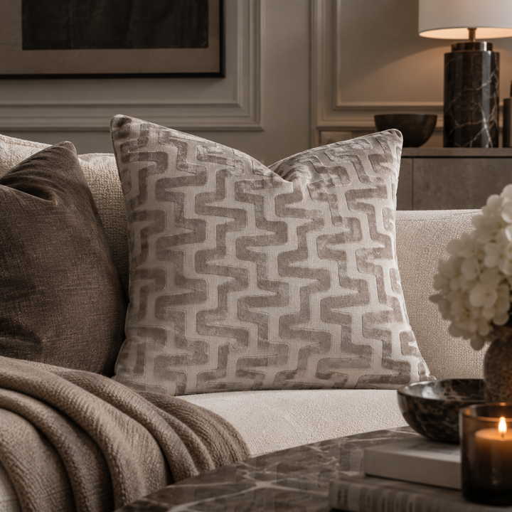 A square throw pillow with a textured geometric chevron pattern in taupe and cream, styled on a light beige sofa in a cozy living room setting.