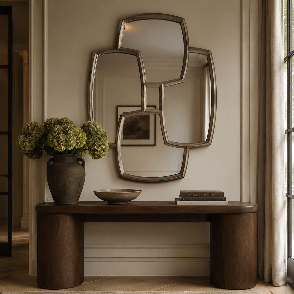 An abstract, multi-panel geometric wall mirror with a brushed silver frame, mounted on a beige wall above a dark wood console table decorated with a vase of green hydrangeas and books.