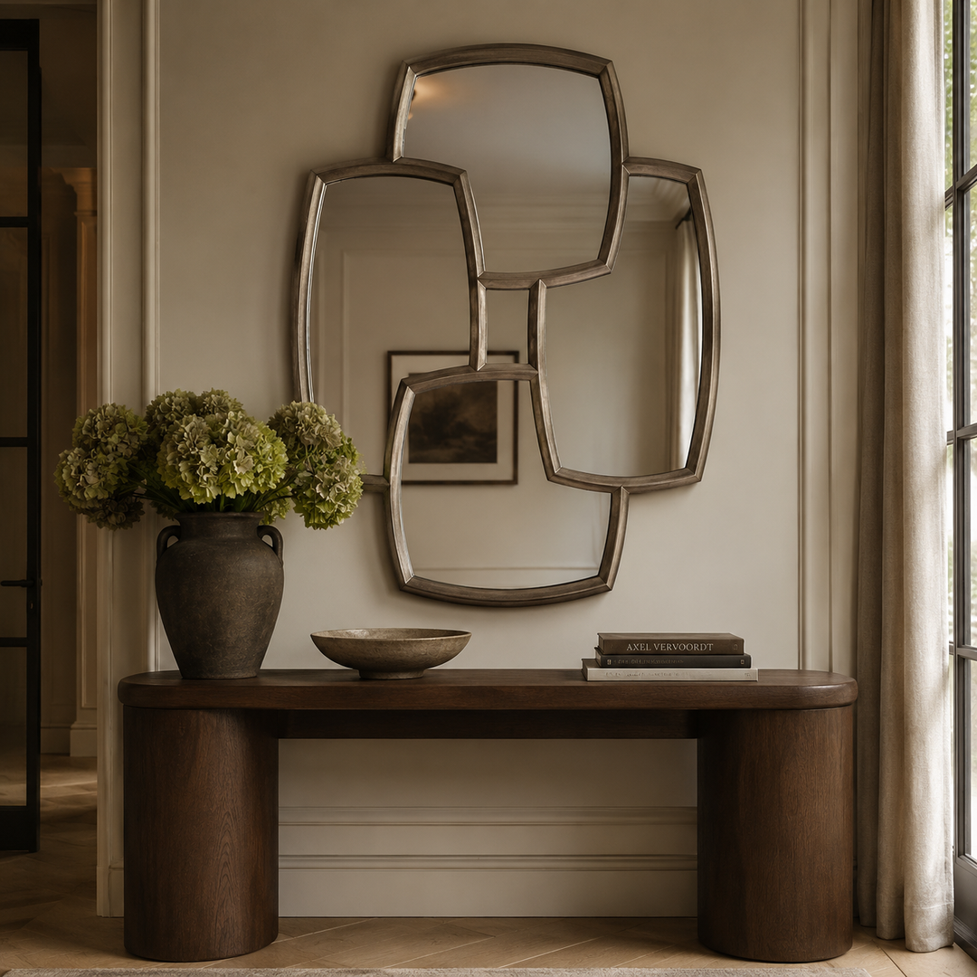 An abstract, multi-panel geometric wall mirror with a brushed silver frame, mounted on a beige wall above a dark wood console table decorated with a vase of green hydrangeas and books.
