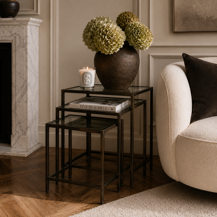 Three nested square side tables with black metal frames and glass tops, styled in a living room next to a cream sofa and a fireplace. The top table holds a dark brown ceramic vase with green hydrangeas and a white candle.