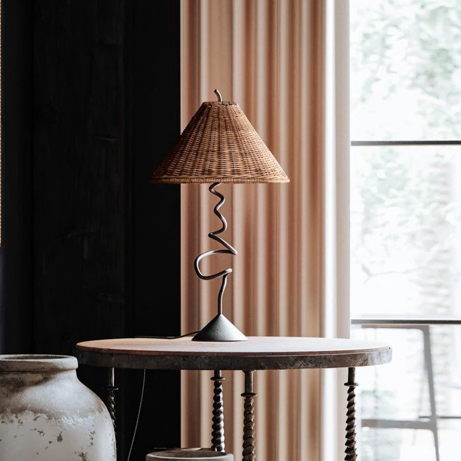 Matte black table lamp with wavy base and rattan shade on a rustic side table against a window and curtain backdrop