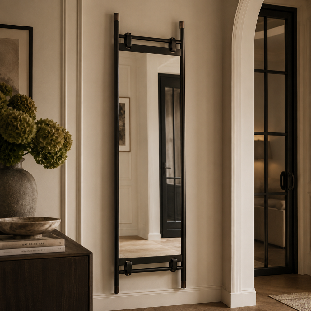 A tall, narrow black metal wall mirror with integrated towel bars, mounted on a beige wall in a styled interior hallway.