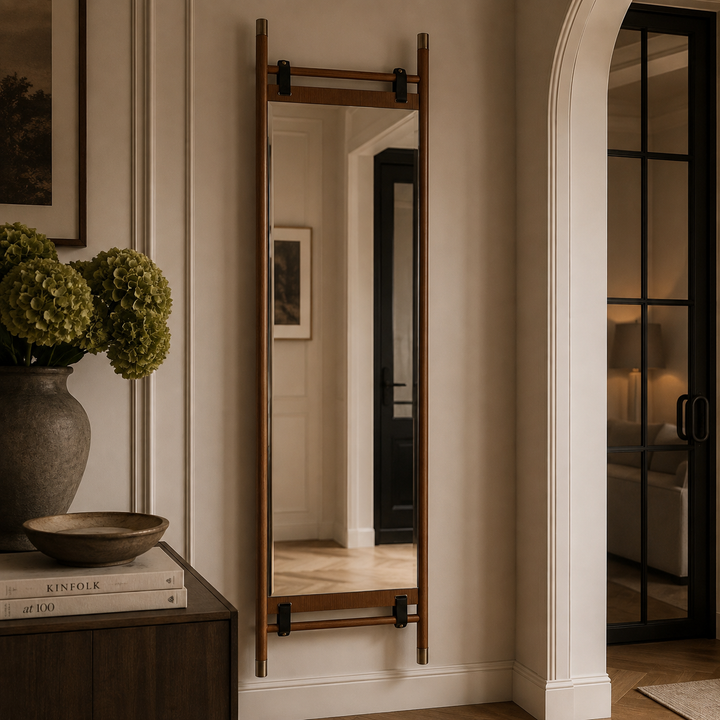 A tall, narrow full-length floor mirror with a warm brown wooden frame and black metal hardware, mounted on a beige wall in a styled interior hallway.