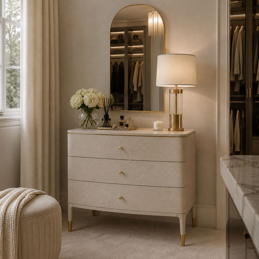 A cream-colored three-drawer dresser with gold legs and knobs, styled in a luxurious bedroom or dressing area with a matching arched mirror, a lit table lamp, and white flowers.