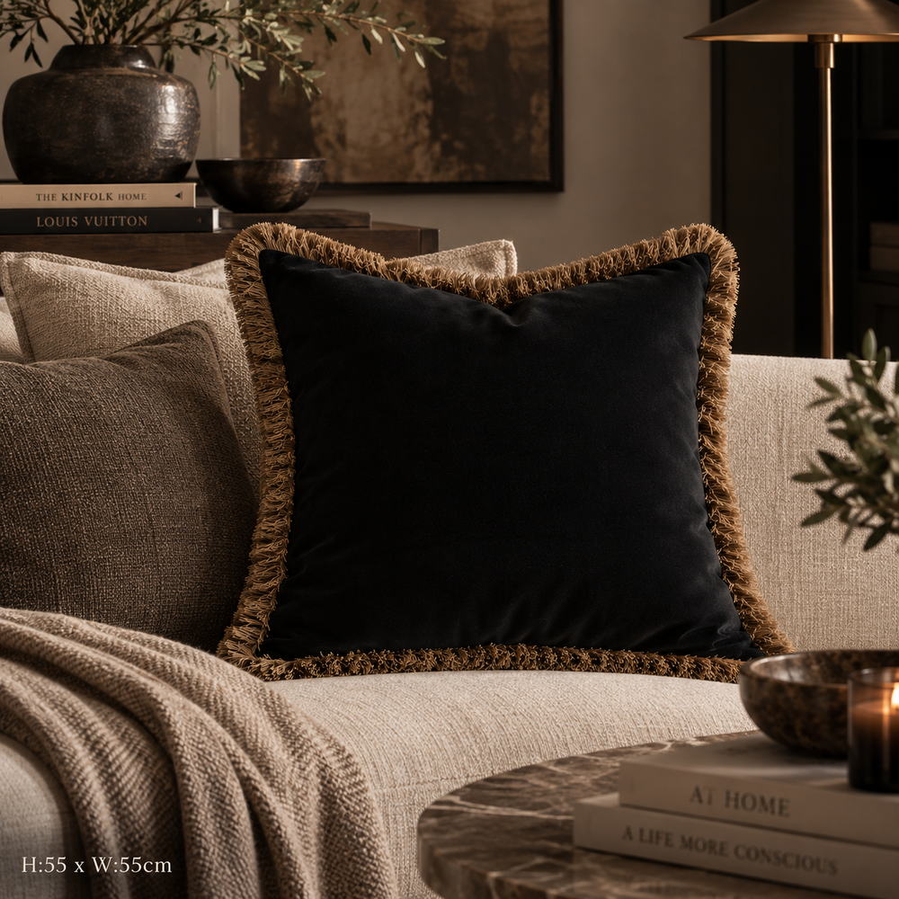 A square black velvet throw pillow with a textured tan fringe border, styled on a beige linen sofa in a warm, cozy living room setting.
