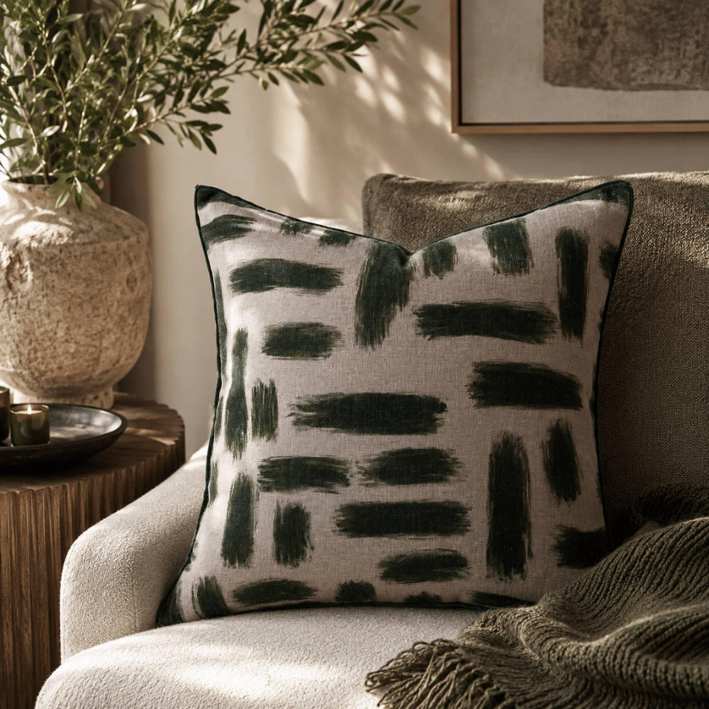 Decorative pillow with black abstract pattern on a beige armchair, with a plant and framed picture in the background.