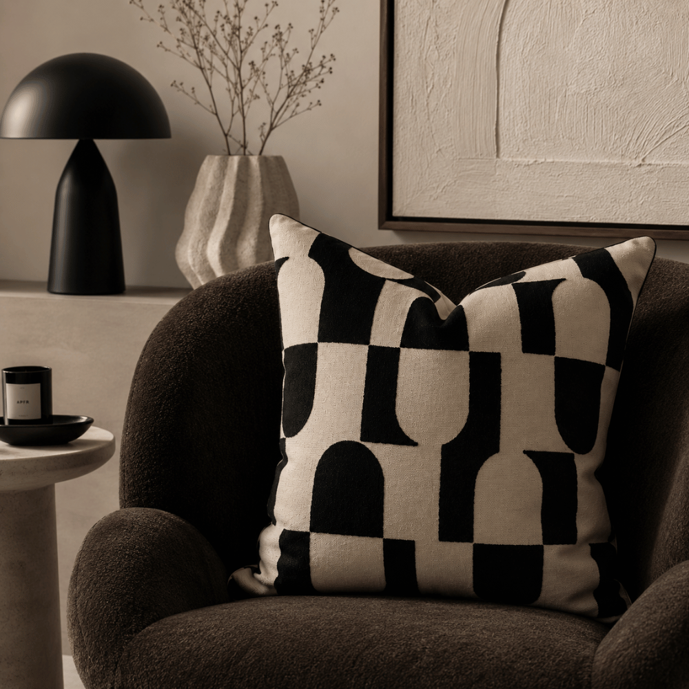 Decorative checkered pillow on a brown armchair with a black lamp and abstract painting in the background.