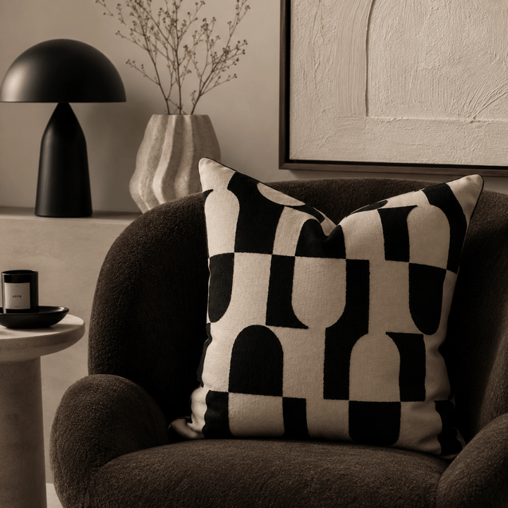 Decorative checkered pillow on a brown armchair with a black lamp and abstract painting in the background.