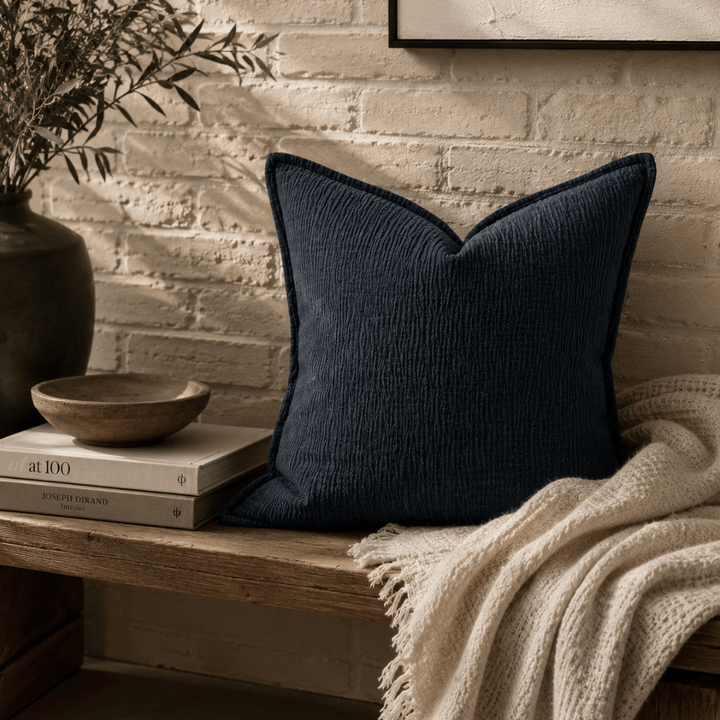 Navy pillow on a wooden bench with books and a plant in the background.