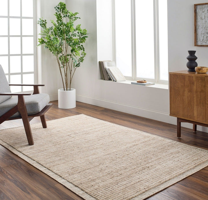 A modern, minimal living room featuring the Yasmin Hand Knotted Rug - Natural, modern furniture, a wooden chair with gray cushion, potted plant, wooden cabinet, and a window seat with books—all against white walls and large windows.